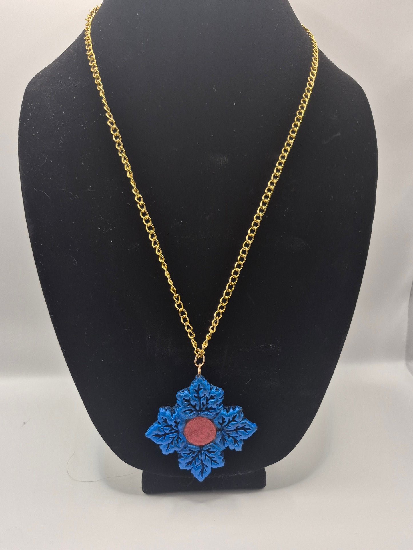 A gold chain necklace with a blue Maple  leaf pendant featuring a central red circular detail, displayed on a black jewelry bust against a white background.