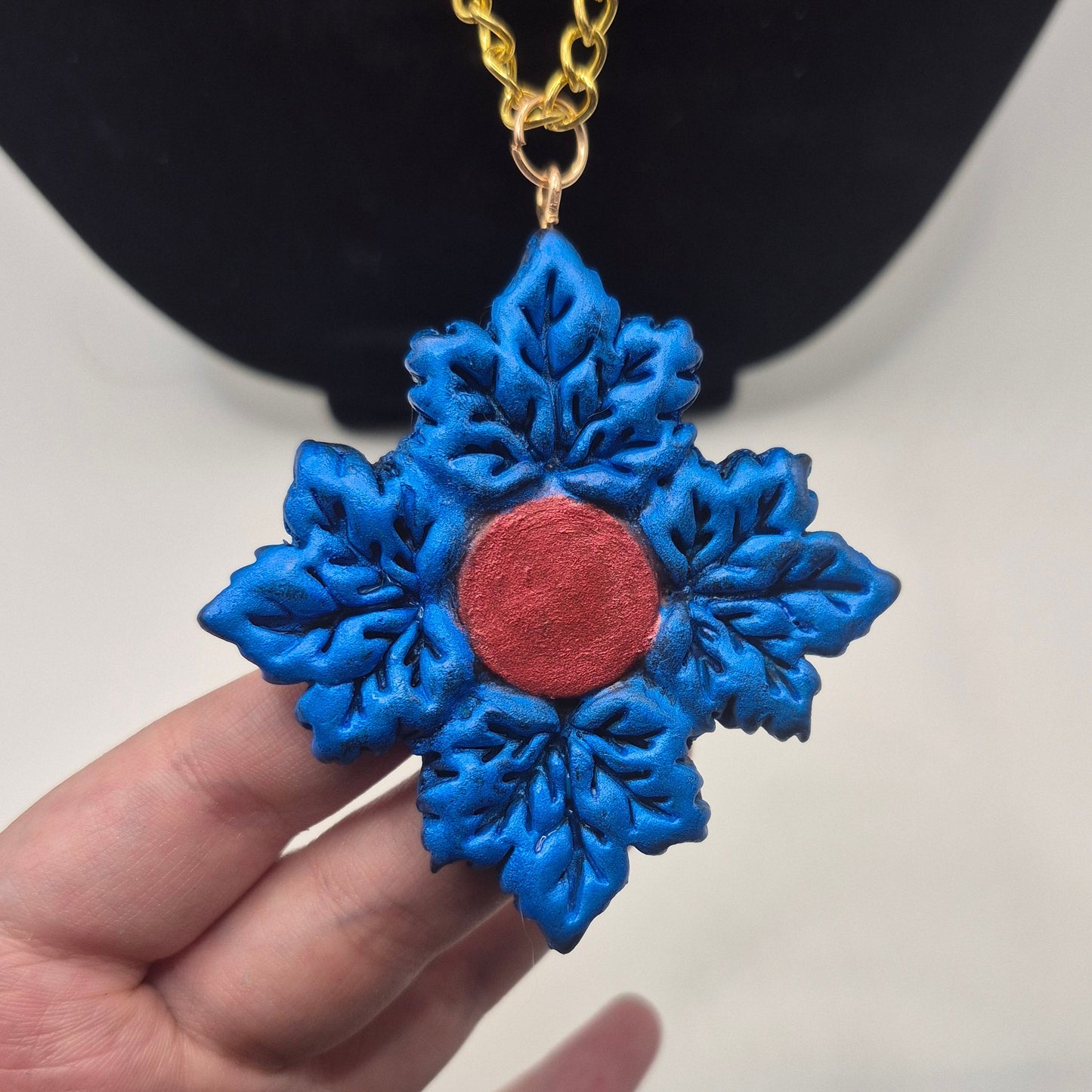 blue maple leaf pendant necklace with a red center and a gold chain, displayed against a black jewelry bust and white background.